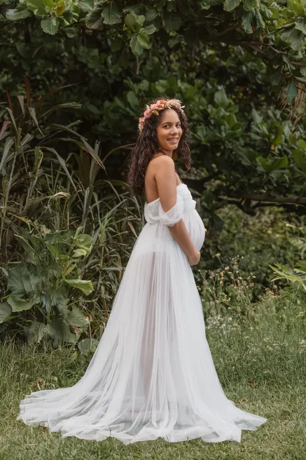 Future maman en longue robe de tulle blanc, couronne de fleurs, se retourne dans l’herbe