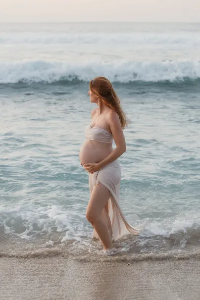 Future maman drapée de blanc marchant dans les vagues, main posée sur son ventre