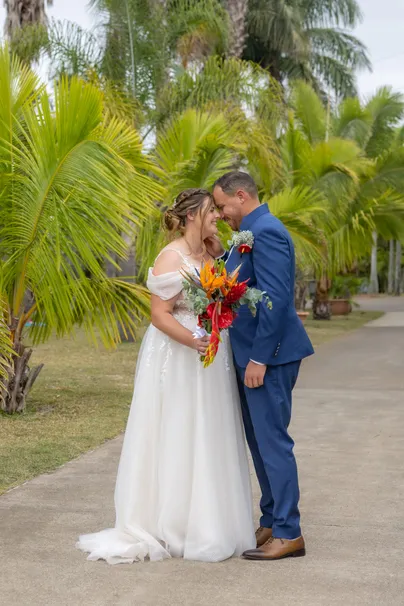 Les mariés front contre front dans une allée bordée de palmiers, bouquet coloré en main