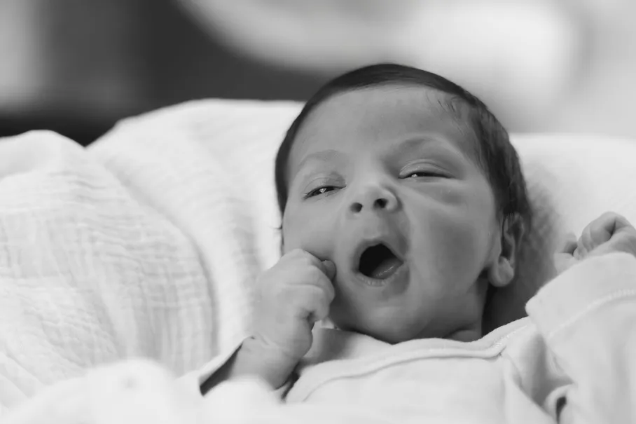 En noir et blanc, le nouveau-né bâille, la main près de la bouche, dans des draps blancs
