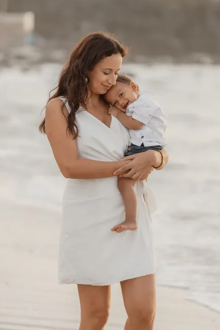 Une mère en robe blanche serre son bébé contre sa joue, au bord des vagues