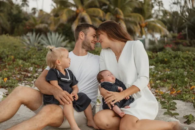 Assis dans le sable sous les palmiers, les parents tiennent leurs deux enfants, le bébé dans les bras de sa mère