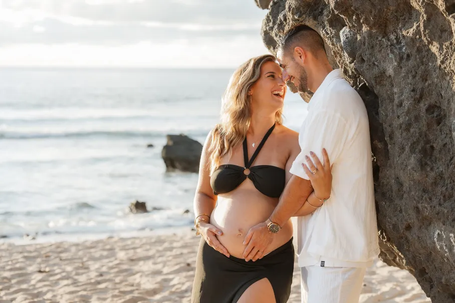 Un couple contre un rocher au bord de la mer, les mains sur le ventre de la future maman