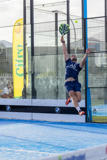 Un joueur de padel saute pour frapper la balle, sur un terrain bleu vu à travers la grille