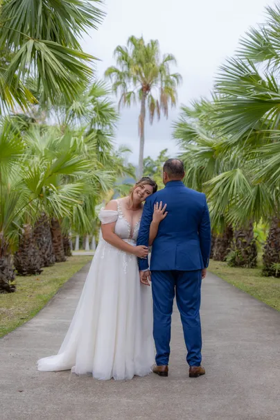 La mariée pose la tête sur l’épaule du marié, vus de dos dans une allée de palmiers