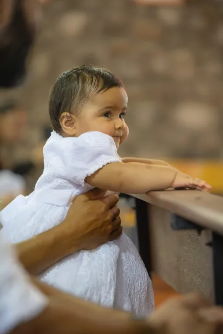 Un bébé en tenue blanche, soutenu par une main, se penche vers le dossier d’un banc d’église
