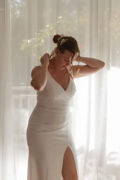 La mariée en robe fendue ajuste ses boucles d’oreilles devant un voilage baigné de lumière