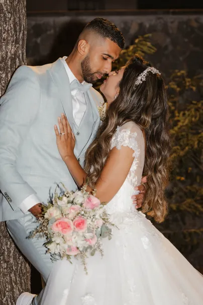 Les mariés s’embrassent contre un tronc d’arbre, la mariée tient un bouquet de roses pâles