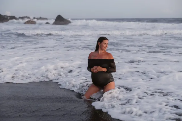 Future maman à genoux dans l’écume, robe noire, sur une plage de sable noir