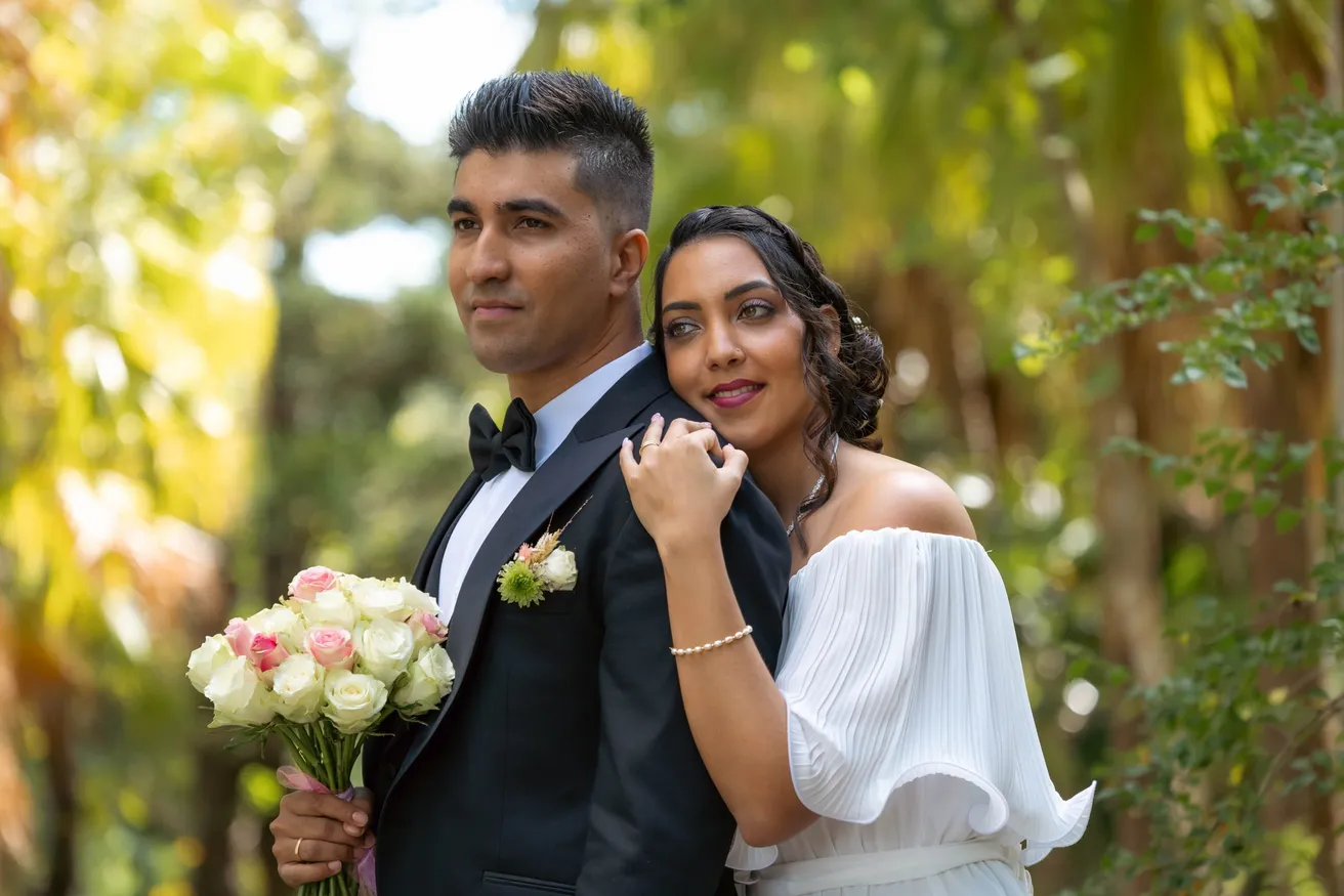 La mariée enlace le marié en smoking par l’épaule, dans la lumière d’un jardin