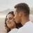 Un couple en séance grossesse assis sur le sable, il l’embrasse sur la joue