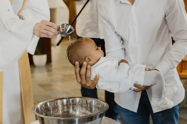 Pendant le baptême, le bébé en blanc est tenu au-dessus de la vasque, l’eau coule sur sa tête