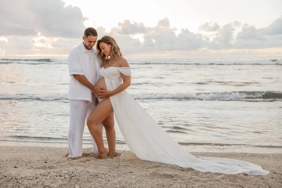 Future maman en robe blanche et son compagnon enlacés sur le sable, face à la mer, la traîne étalée derrière elle
