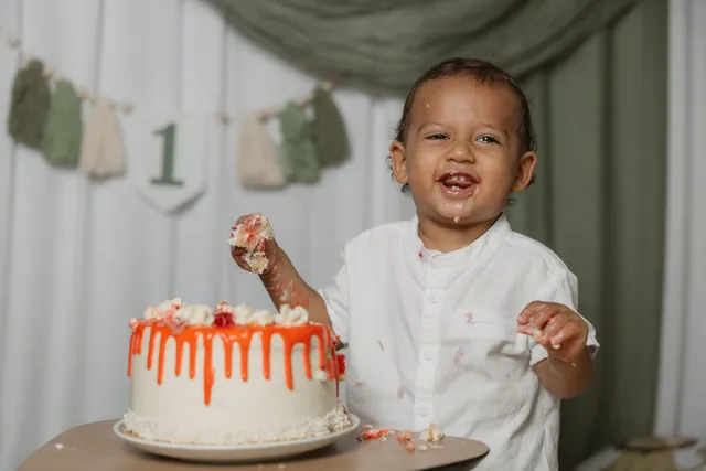 Un petit garçon en chemise blanche éclate de rire, la main pleine de crème, devant son gâteau d’anniversaire orange