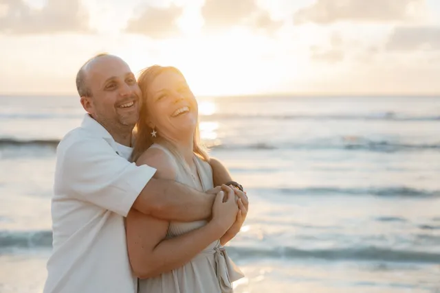 Au coucher du soleil, un homme serre sa compagne contre lui et tous deux éclatent de rire face à la mer