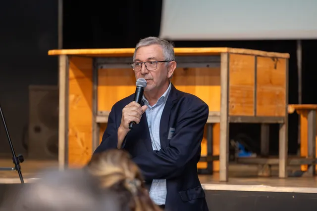 Un homme en costume parle au micro sur la scène d’une salle de conférence