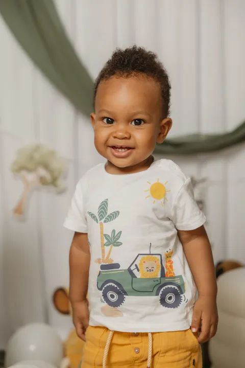 Un petit garçon en tee-shirt imprimé d’une jeep et short moutarde sourit, debout devant le décor safari