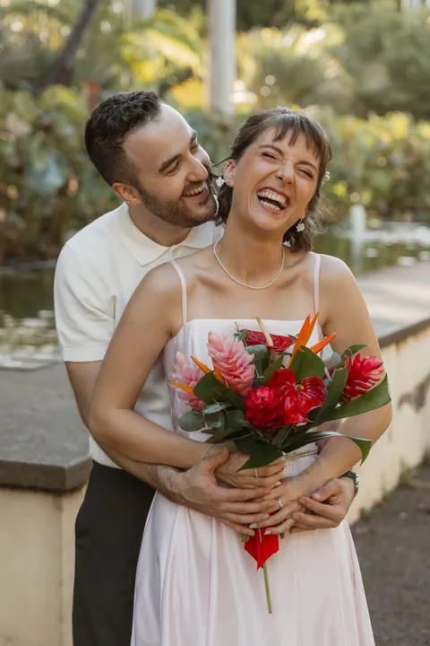 Le marié enlace la mariée par-derrière, elle éclate de rire, un bouquet de fleurs tropicales à la main