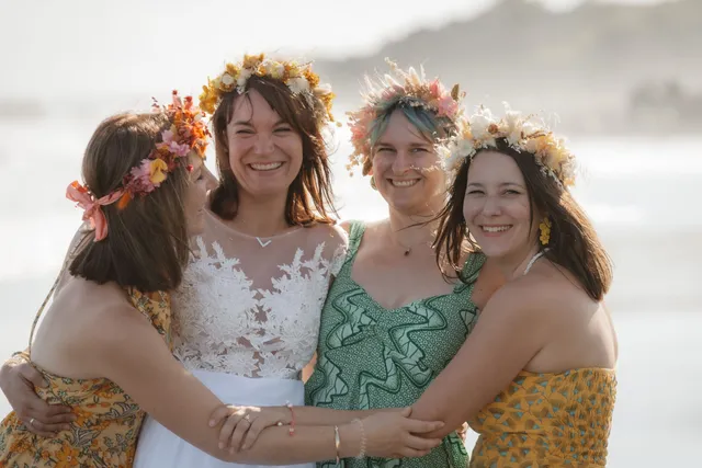 La future mariée en dentelle blanche et ses trois amies, couronnes de fleurs sur la tête, s’enlacent en riant au bord de la mer
