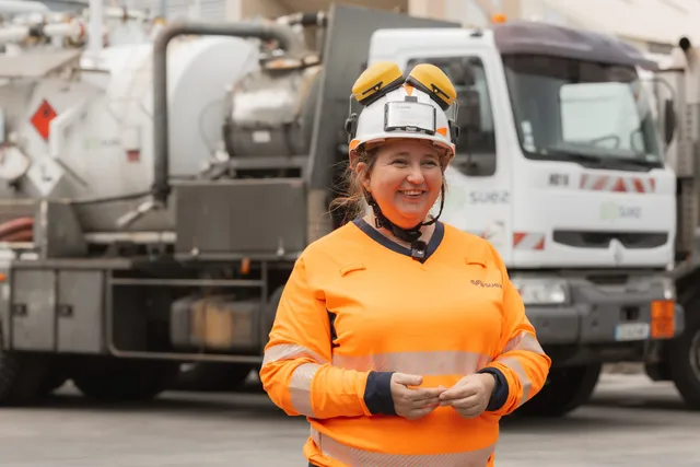 Une salariée en tenue orange et casque blanc sourit, bras croisés, devant un camion-citerne sur un site industriel