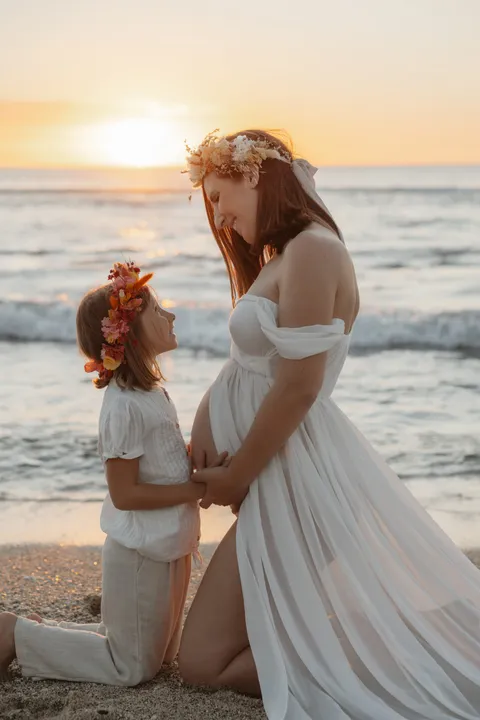 Une petite fille en couronne de fleurs, à genoux face au ventre rond de sa maman, au soleil couchant