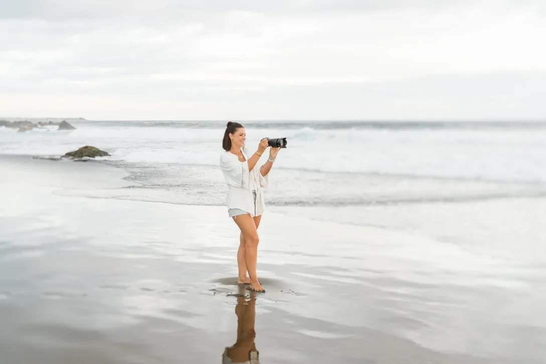 Lucile Boissé, pieds nus sur le sable mouillé, sourit en visant avec son appareil photo face aux vagues