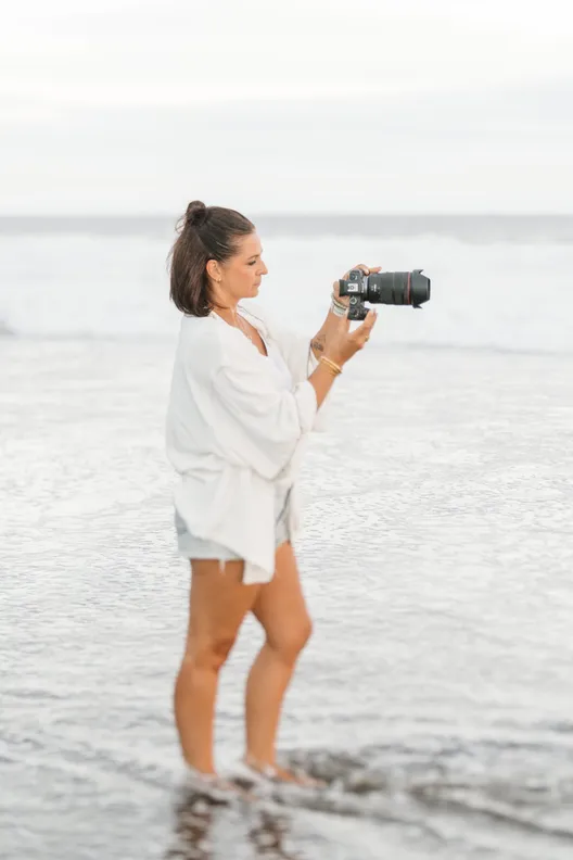 Lucile Boissé, en chemise blanche et les pieds dans l’eau, vise avec son appareil photo au bord des vagues